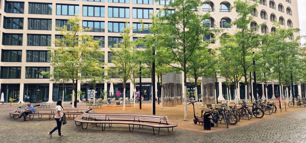 Urban park with benches and trees, featuring modern architecture in the background. The area is designed for relaxation and social gatherings, showcasing greenery amidst a cityscape.
