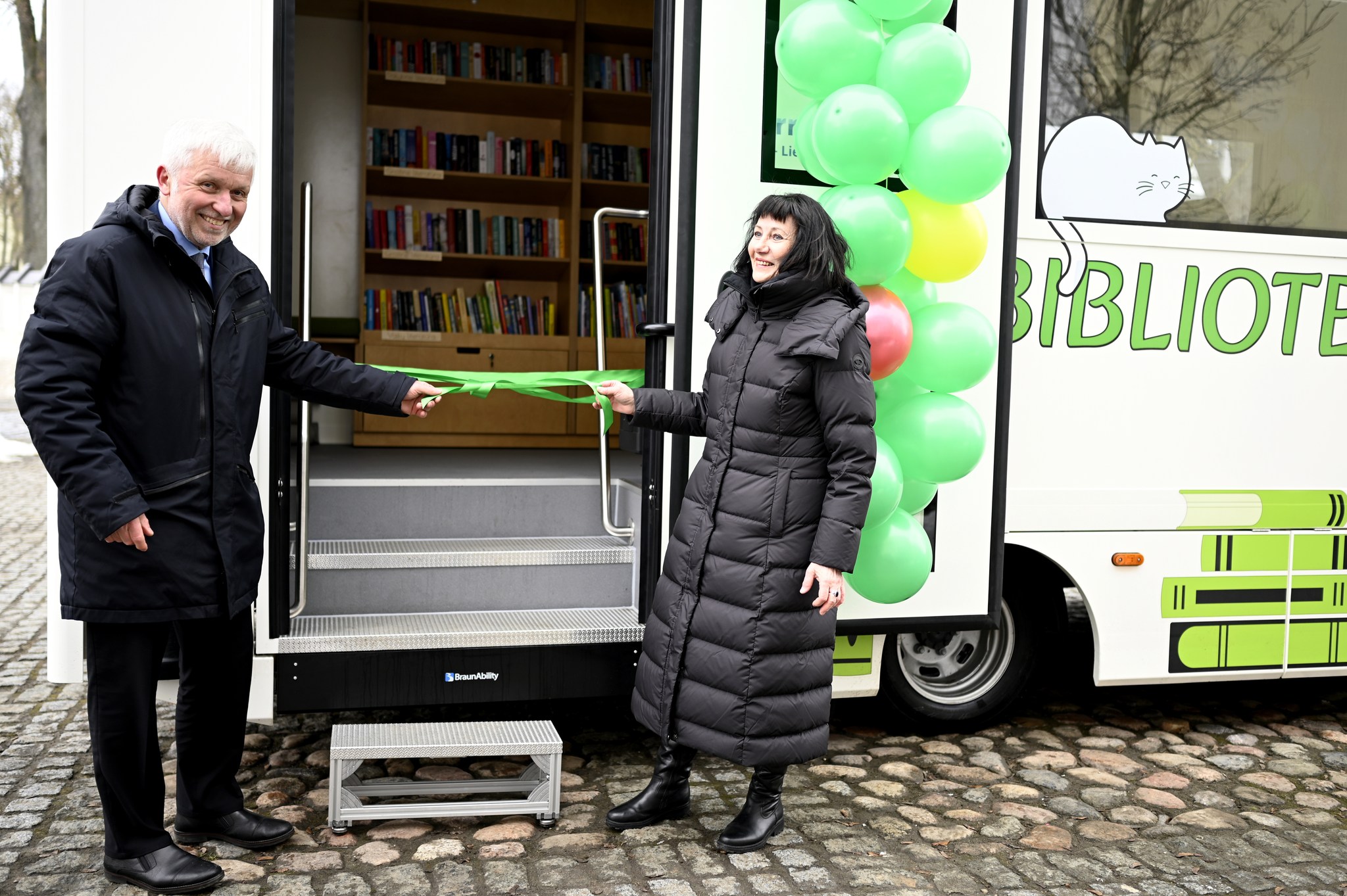 A man and a woman stand outside a mobile library, preparing to cut a green ribbon to officially inaugurate the bookmobile. The vehicle is decorated with colorful balloons and features shelves filled with books visible inside. A man and a woman stand outside a mobile library, preparing to cut a green ribbon to officially inaugurate the bookmobile. The vehicle is decorated with colorful balloons and features shelves filled with books visible inside.