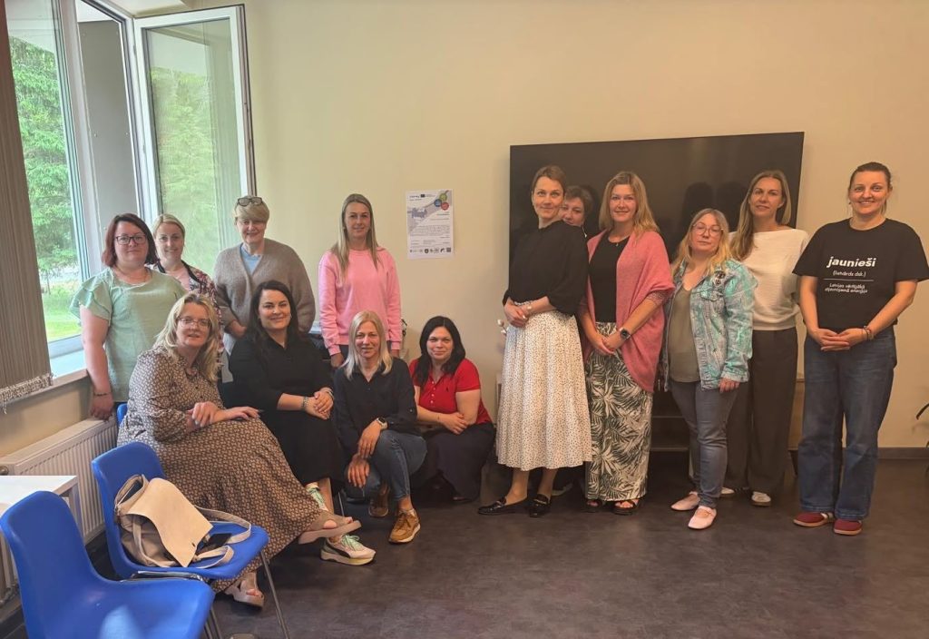 Group photo of women participating in a community event, standing and sitting in a bright room with large windows. The women are dressed in a variety of casual and semi-formal attire, showcasing diversity in styles. A poster is visible on the wall, indicating the event theme or organization. Group photo of women participating in a community event, standing and sitting in a bright room with large windows. The women are dressed in a variety of casual and semi-formal attire, showcasing diversity in styles. A poster is visible on the wall, indicating the event theme or organization.