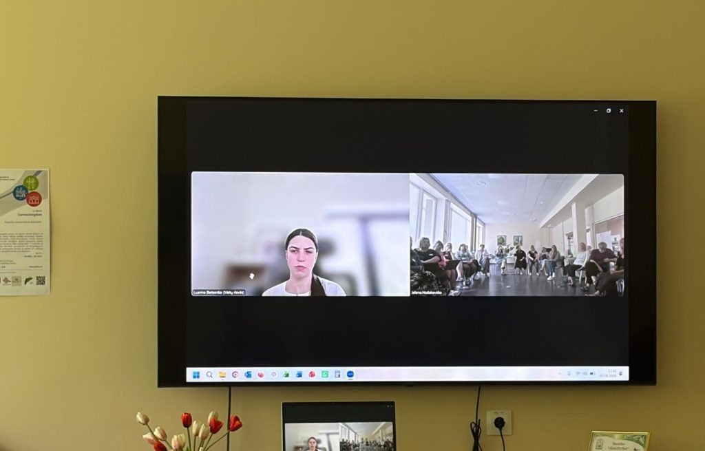 Video conference session displayed on a large screen, featuring a speaker and a group of participants seated in a well-lit room. A smaller screen at the bottom shows the speaker's image. The setting includes decorative elements, such as flowers, and informational materials on the wall. Video conference session displayed on a large screen, featuring a speaker and a group of participants seated in a well-lit room. A smaller screen at the bottom shows the speaker's image. The setting includes decorative elements, such as flowers, and informational materials on the wall.