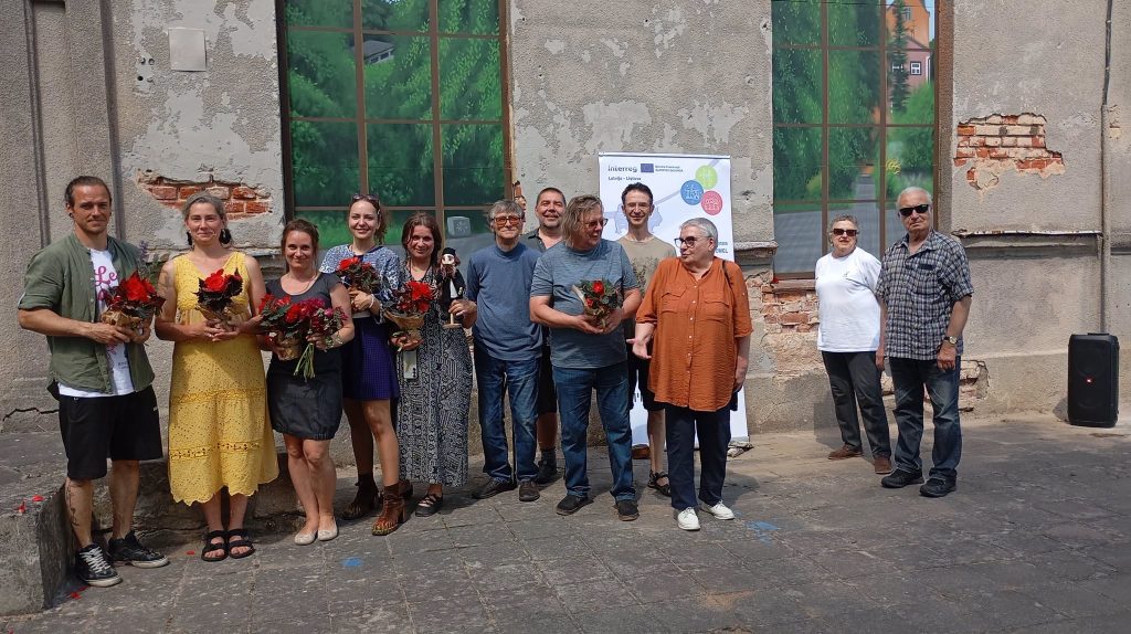 Group photo of diverse individuals holding flower bouquets, standing in front of a weathered wall with large windows and a colorful mural. The setting appears to be an outdoor event, possibly a celebration or community gathering, highlighting collaboration and cultural engagement.