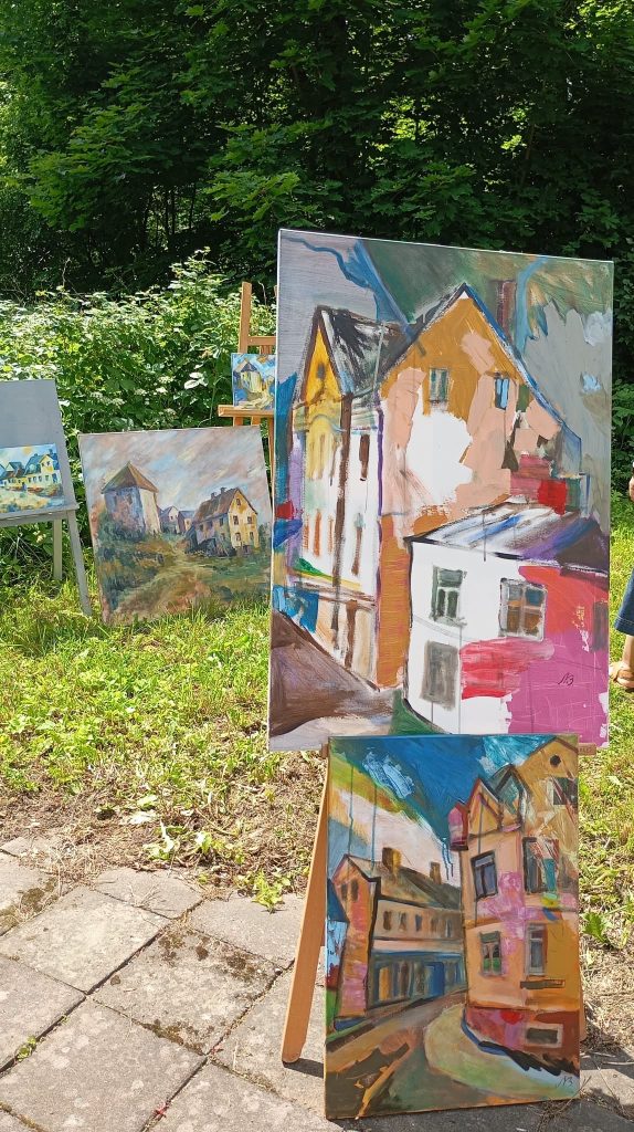 Outdoor art display featuring colorful paintings of buildings and landscapes, with multiple easels showcasing various artistic styles. Lush greenery surrounds the scene, providing a vibrant backdrop for the artworks.