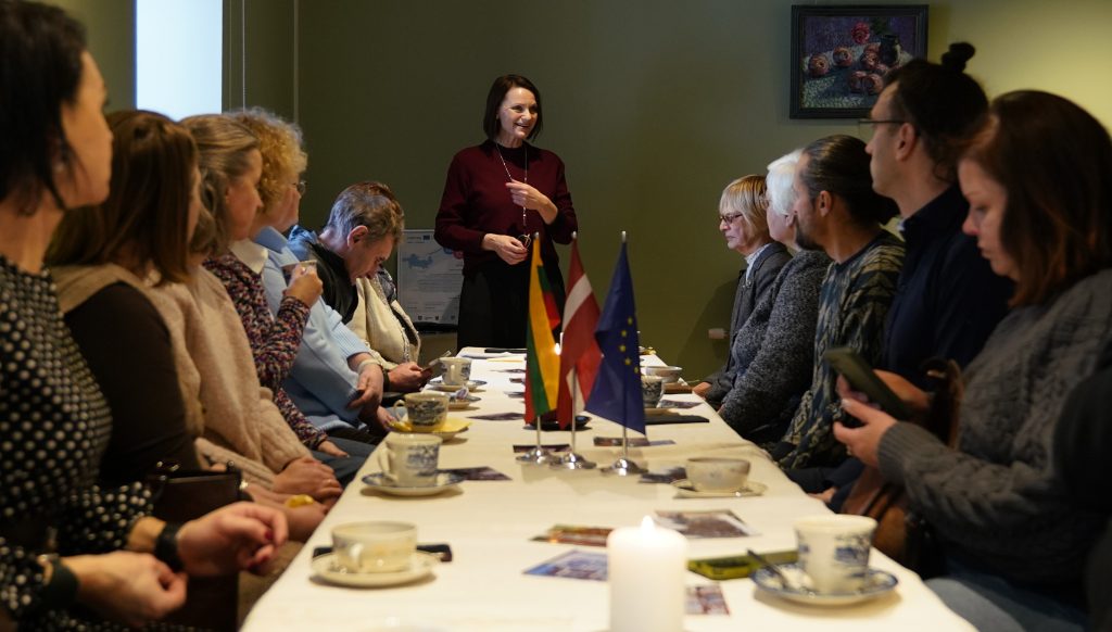 A group of people seated around a table with cups and flags from Germany and the European Union, engaged in a discussion led by a woman standing at the head of the table. The setting appears to be a formal meeting or conference.