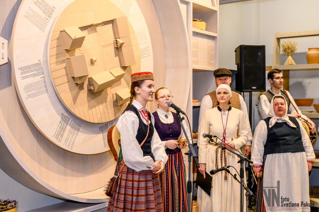 Group of traditional performers in folk costumes singing at a cultural event, with a decorative backdrop featuring a circular design. The setting highlights the importance of heritage and community in the celebration.