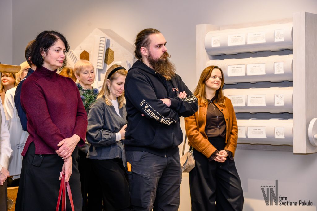 Group of people attending an art exhibition, with diverse expressions and styles, standing in front of a display featuring architectural models and designs.