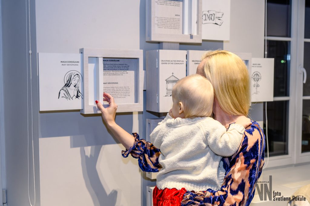 A mother holds her baby while interacting with an art installation featuring illustrated panels on a gallery wall, promoting family engagement in the arts.