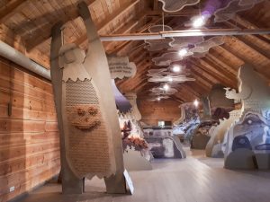 Interior view of a wooden exhibition space featuring large, artistic displays, including a prominent figure with a smiling face made of wooden pegs, showcasing interactive elements and nature-themed illustrations on the walls.