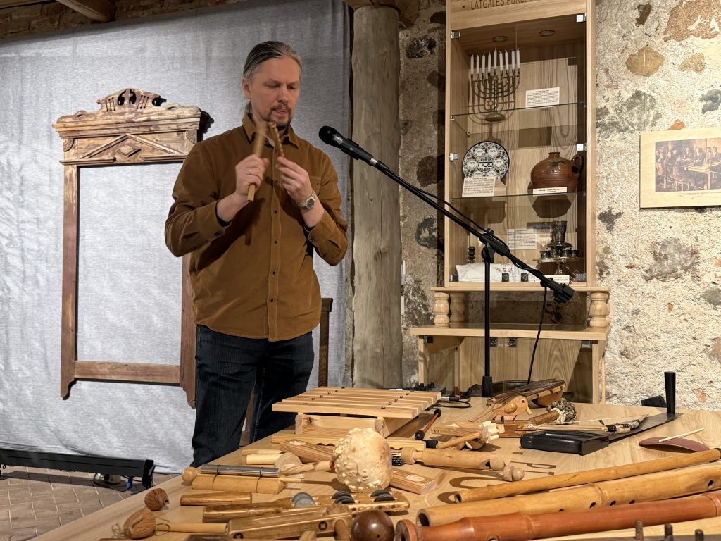 Man playing a wooden flute while standing in front of a table filled with handcrafted wooden instruments and artifacts, with a microphone and display case in the background.