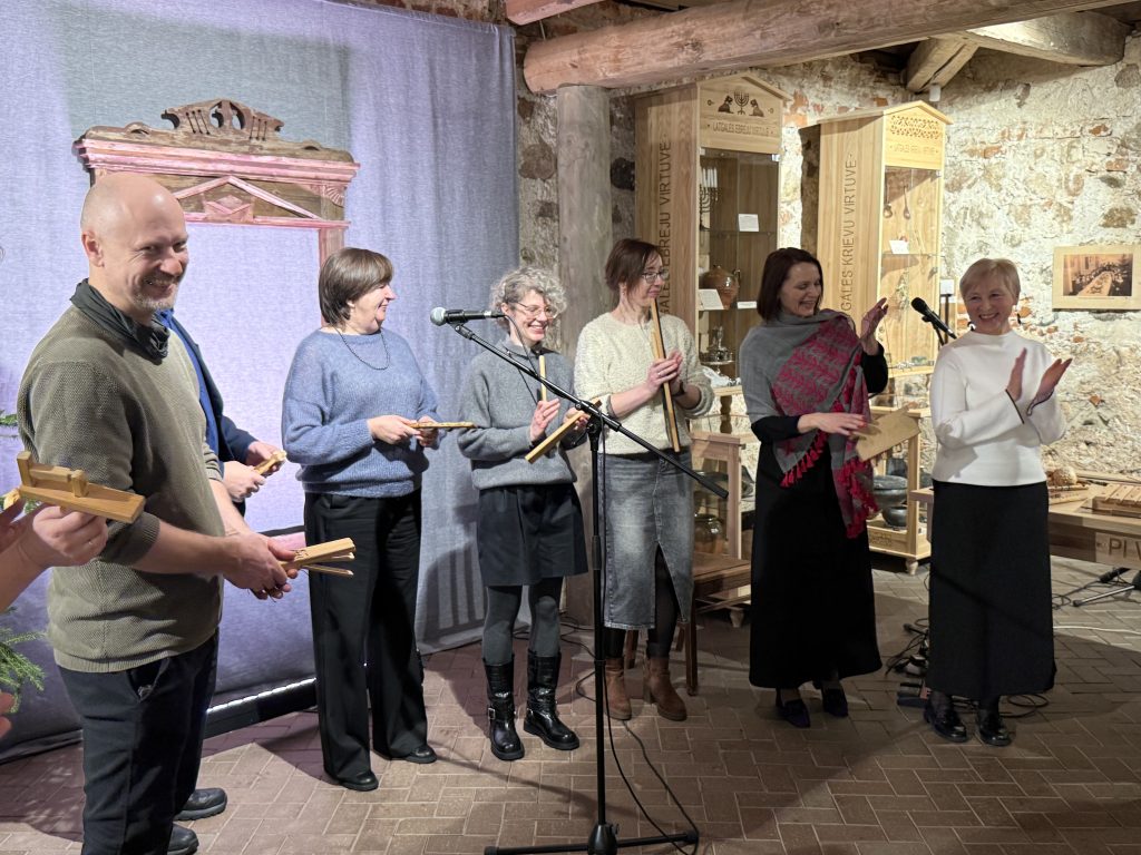 Group of six performers on stage holding instruments and singing during a cultural event in a cozy indoor setting, featuring wooden decor and soft lighting.
