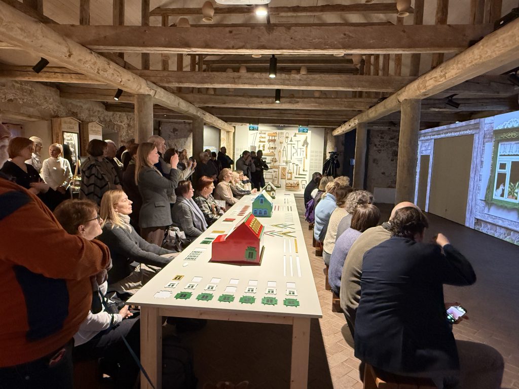 Audience attending a presentation in a rustic venue, seated at a long table with green models, while a speaker presents on a screen in the background.
