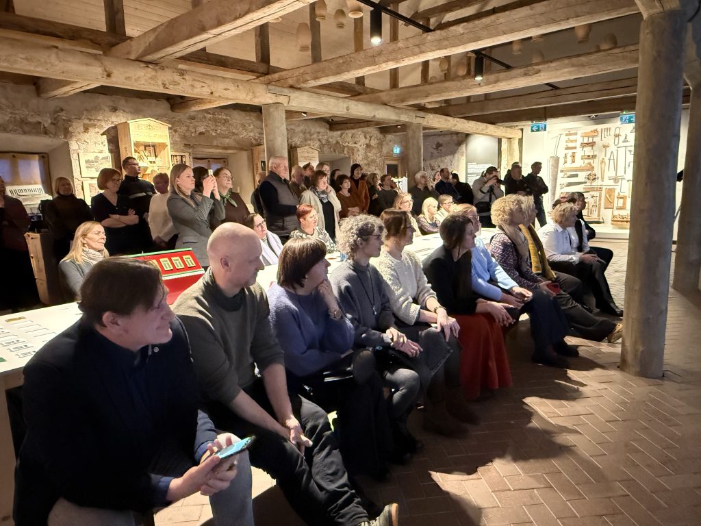 Audience seated in a rustic venue, engaged in a presentation or event, with a diverse group of individuals and a warm, inviting atmosphere.