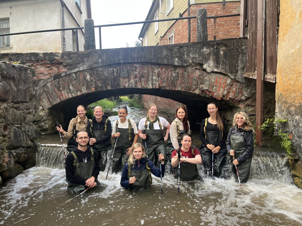 Group of ten individuals wearing waders standing in a stream under a brick arch bridge, engaged in a water-based activity, surrounded by historical buildings and lush greenery.