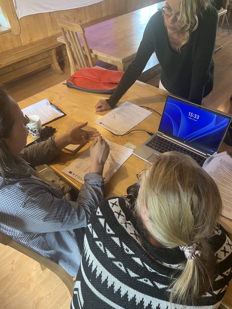Group collaboration in a cozy wooden room, featuring three individuals engaged in discussion around a table with documents and a laptop displaying the time. The setting suggests a productive meeting or workshop atmosphere.