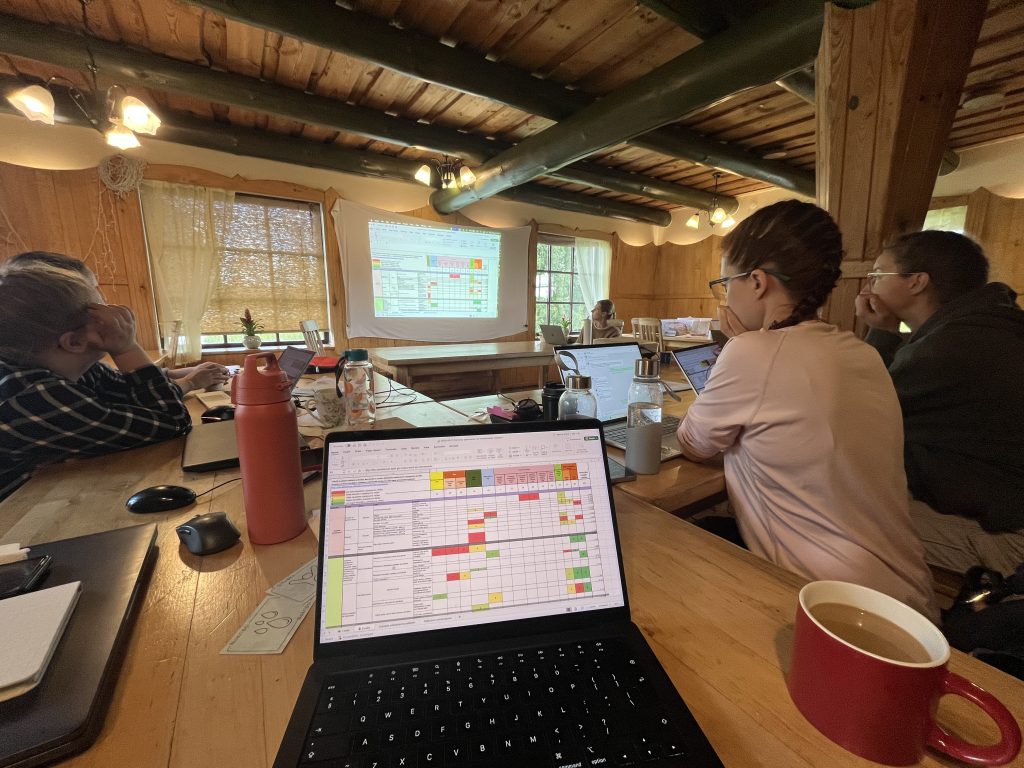 Group meeting in a wooden conference room, featuring individuals engaged in discussion while reviewing a presentation projected on a screen. A laptop displaying a colorful spreadsheet is in the foreground, alongside a water bottle and a coffee cup.