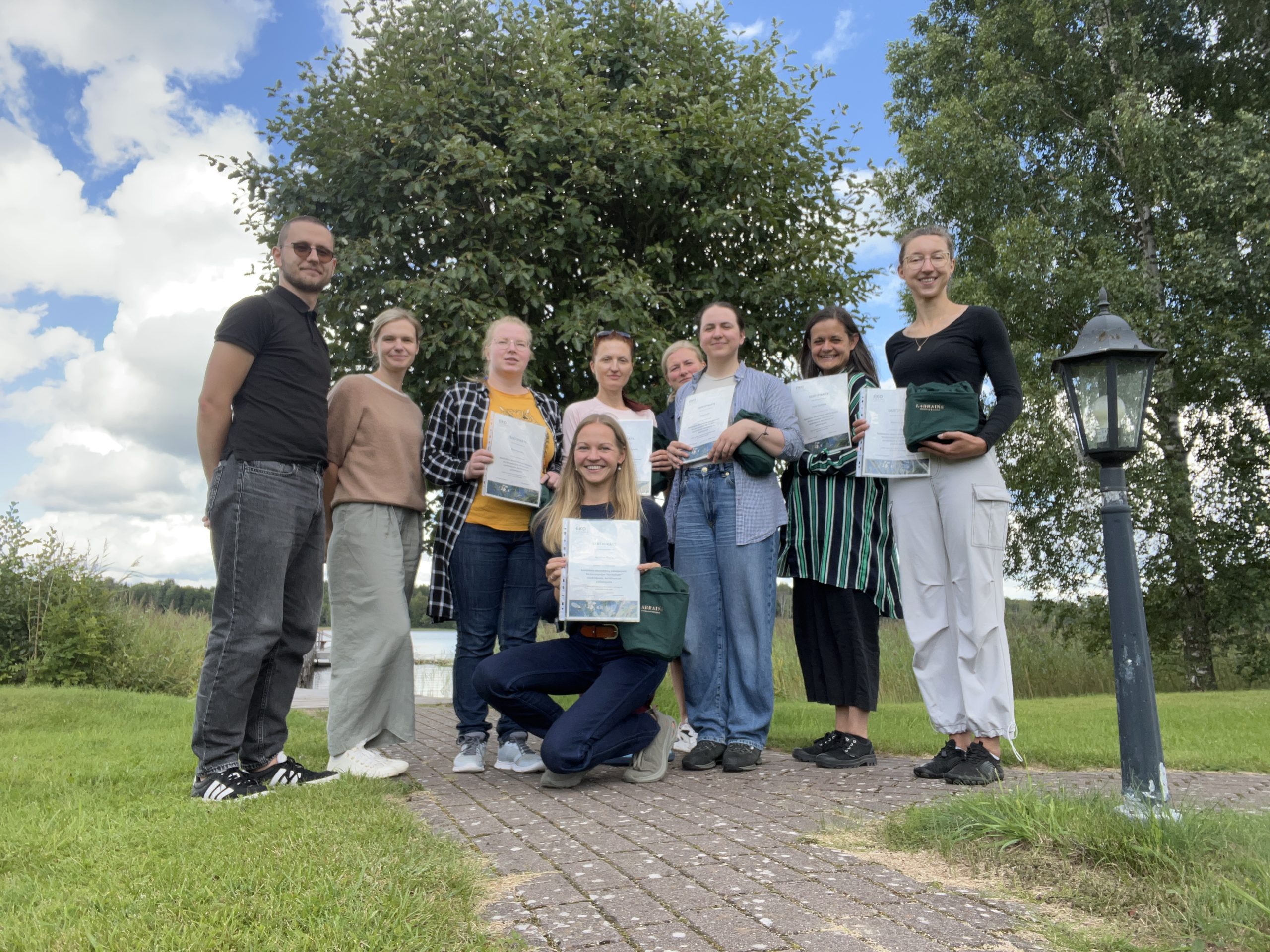 Group photo of eight individuals holding certificates, celebrating a successful completion of a training program outdoors with a scenic background of trees and a cloudy sky.