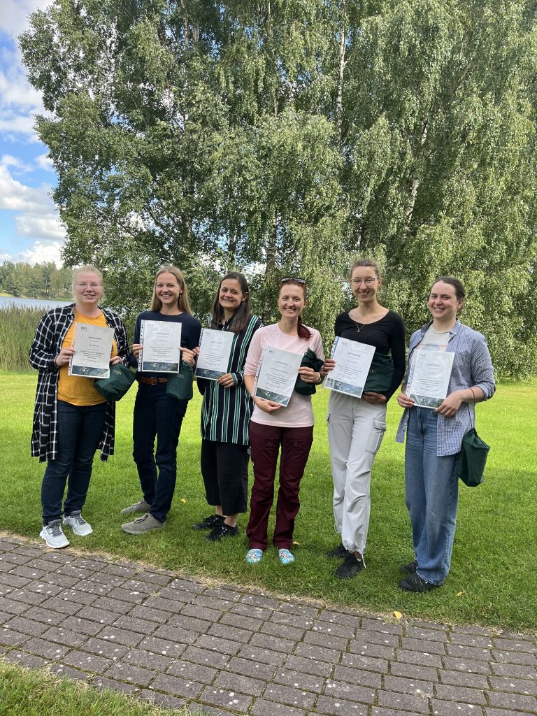 Group of six women proudly holding certificates outdoors, surrounded by greenery and a scenic background, celebrating their achievements.