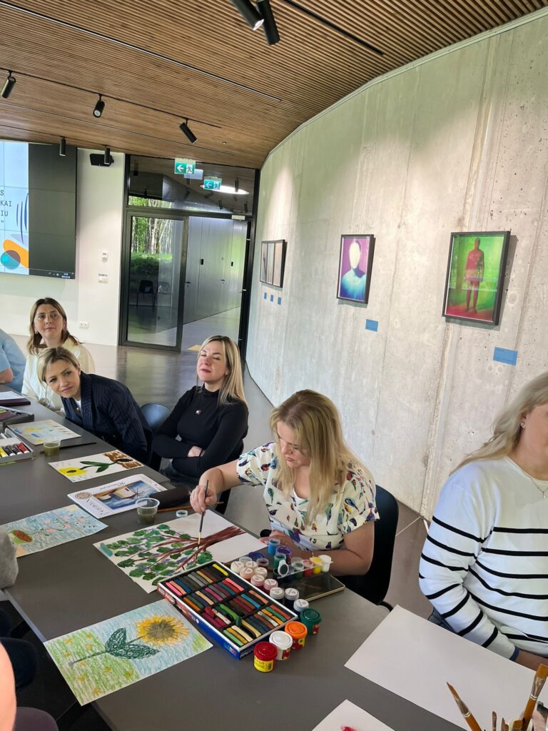 Group of people engaged in a creative workshop, with one participant painting at a table surrounded by art supplies and colorful artworks displayed on the walls. Group of people engaged in a creative workshop, with one participant painting at a table surrounded by art supplies and colorful artworks displayed on the walls.