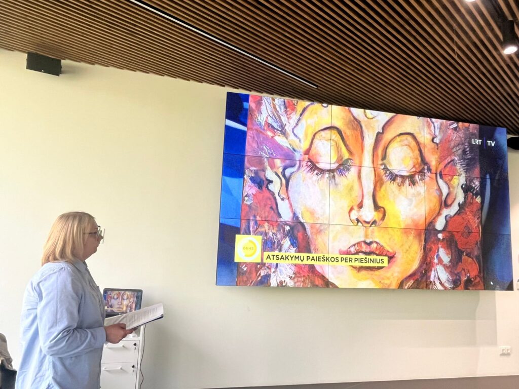 A woman with blonde hair stands beside a large screen displaying a colorful artistic image of a face, while holding a document. The screen features text in Lithuanian, indicating a presentation context. The setting has modern architectural elements with wooden ceiling panels. A woman with blonde hair stands beside a large screen displaying a colorful artistic image of a face, while holding a document. The screen features text in Lithuanian, indicating a presentation context. The setting has modern architectural elements with wooden ceiling panels.