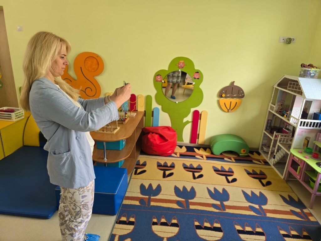 A woman taking a photo in a colorful children's playroom featuring vibrant wall decorations, a patterned rug, and various play items like bean bags and toys. A woman taking a photo in a colorful children's playroom featuring vibrant wall decorations, a patterned rug, and various play items like bean bags and toys.