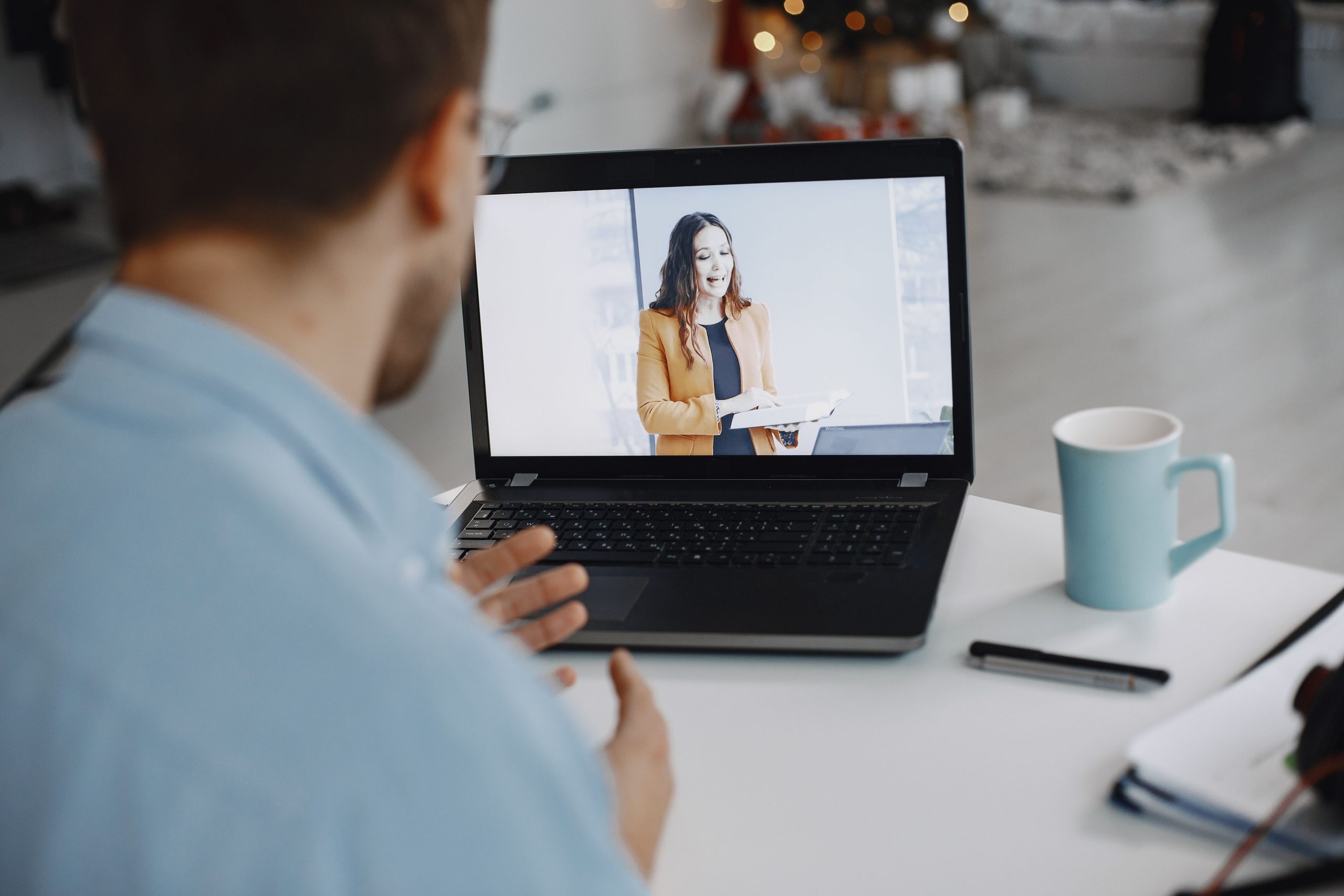 A person sitting at a desk engages in a video call on a laptop, watching a woman in a professional outfit presenting. A blue mug and a pen are visible on the table, suggesting a home office setting.