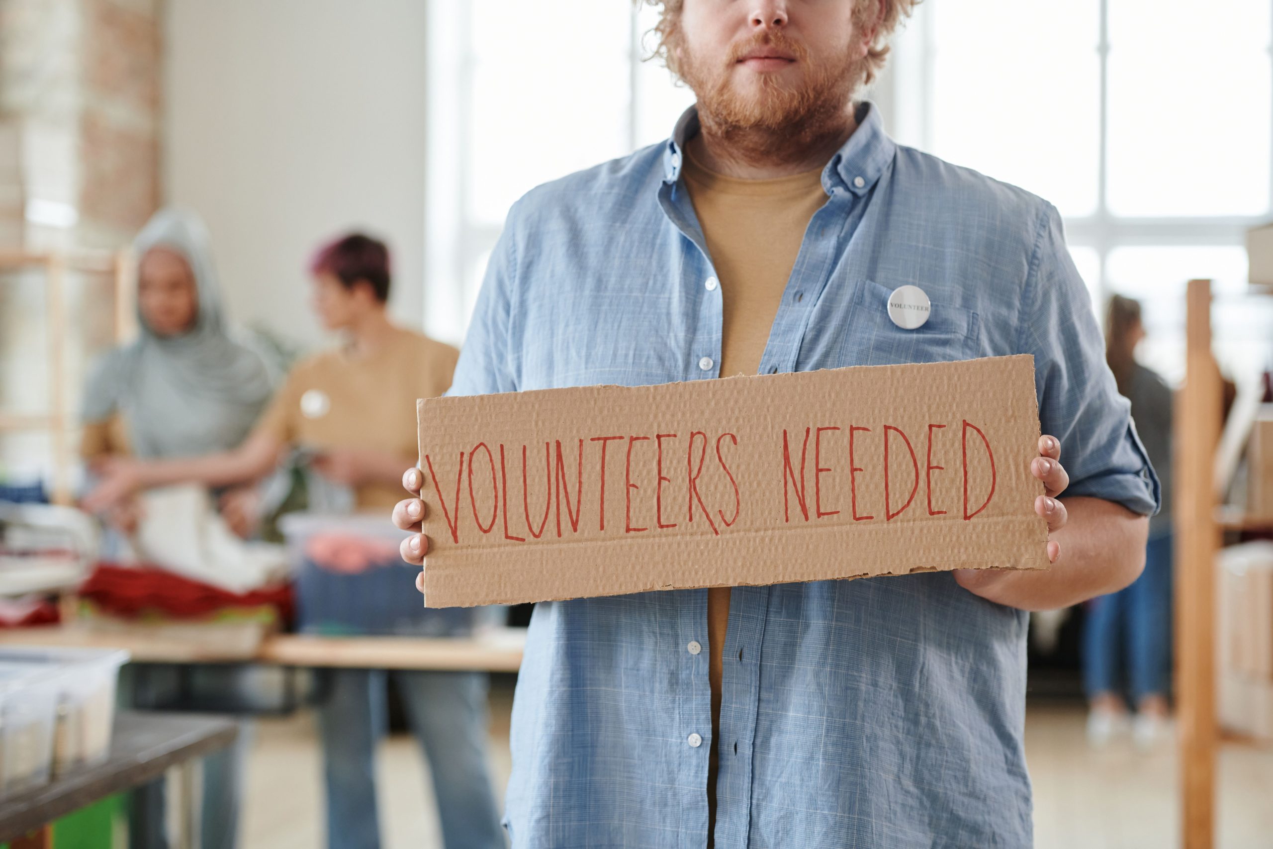 Person holding a cardboard sign that reads "Volunteers Needed" in a community center, with other volunteers engaged in activities in the background, emphasizing the importance of community involvement and volunteer work. Person holding a cardboard sign that reads "Volunteers Needed" in a community center, with other volunteers engaged in activities in the background, emphasizing the importance of community involvement and volunteer work.