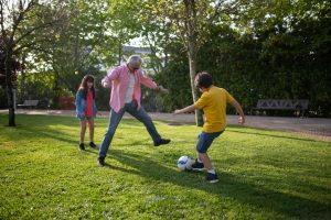 A grandfather playing soccer with his two grandchildren in a sunny park, surrounded by green grass and trees, promoting family bonding and outdoor activities.