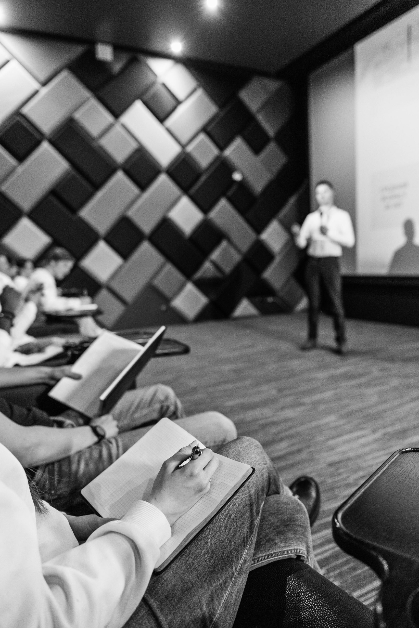 Audience taking notes during a presentation in a modern lecture hall, featuring a speaker in the background and a textured wall design.