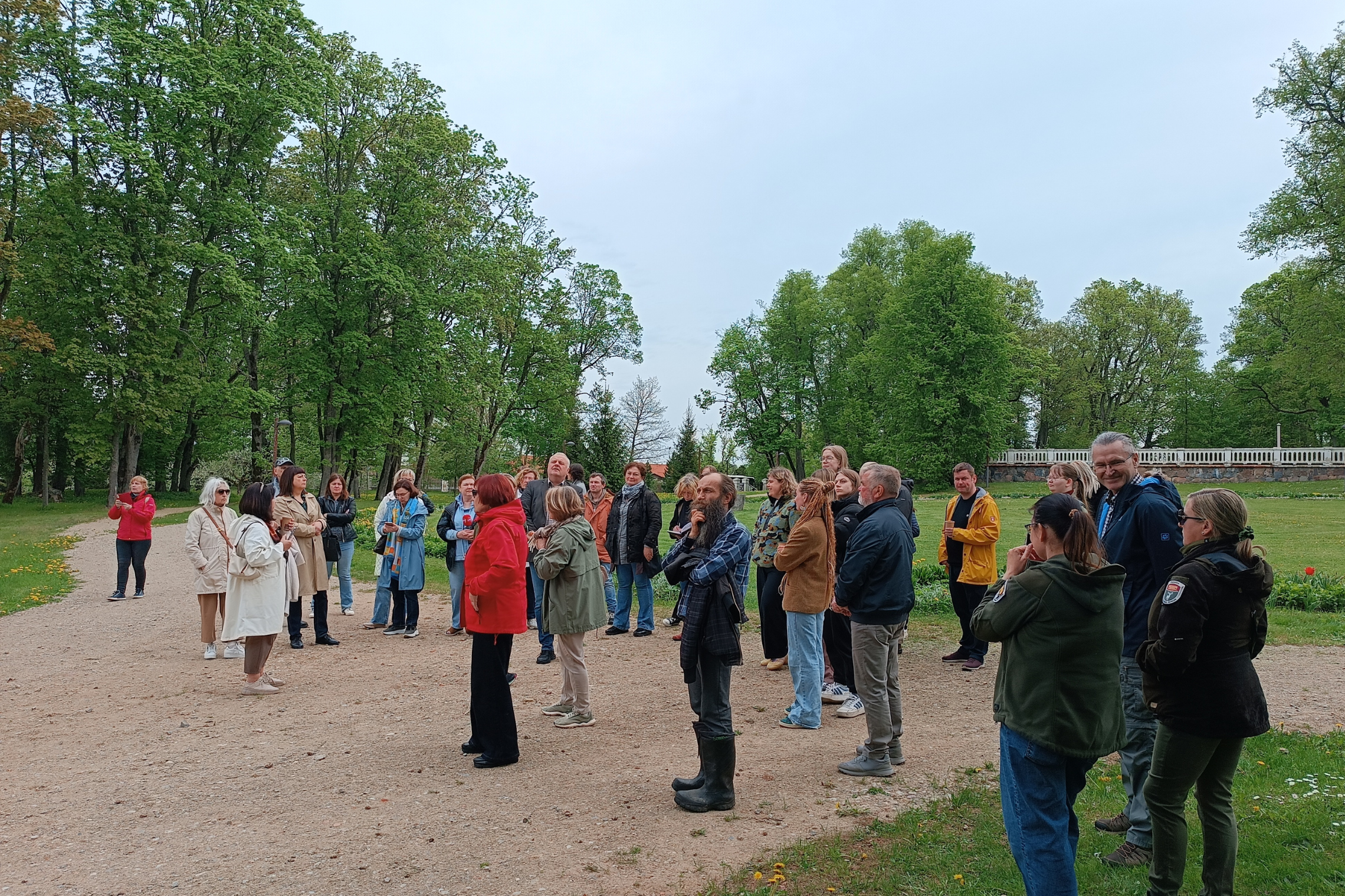 Group of people gathered in a park, engaged in conversation, surrounded by lush greenery and trees, during a daytime event.