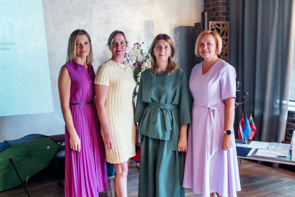 Four women standing together in a stylish indoor setting, dressed in colorful dresses. The background features a projector screen and decorative elements, including flags and floral arrangements. The image captures a moment of collaboration and empowerment among professionals. Four women standing together in a stylish indoor setting, dressed in colorful dresses. The background features a projector screen and decorative elements, including flags and floral arrangements. The image captures a moment of collaboration and empowerment among professionals.
