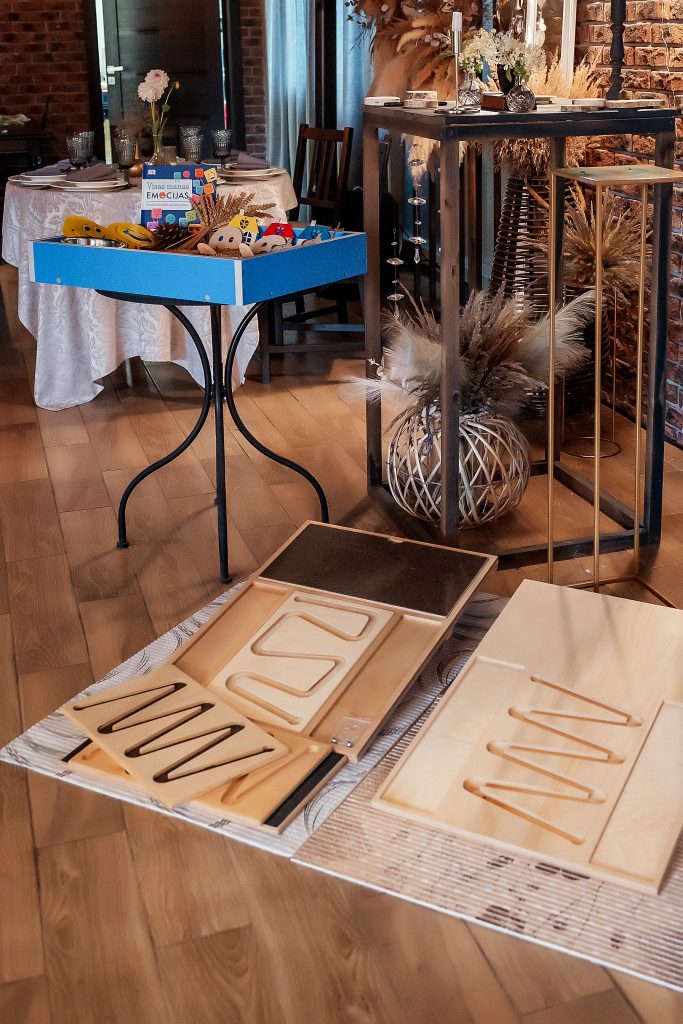 Indoor setting featuring a wooden table with a blue box filled with colorful toys and learning materials. Nearby, two wooden boards with unique patterns are displayed on a woven mat, showcasing a playful and educational environment. Natural decor elements, including dried plants, enhance the cozy atmosphere. Indoor setting featuring a wooden table with a blue box filled with colorful toys and learning materials. Nearby, two wooden boards with unique patterns are displayed on a woven mat, showcasing a playful and educational environment. Natural decor elements, including dried plants, enhance the cozy atmosphere.