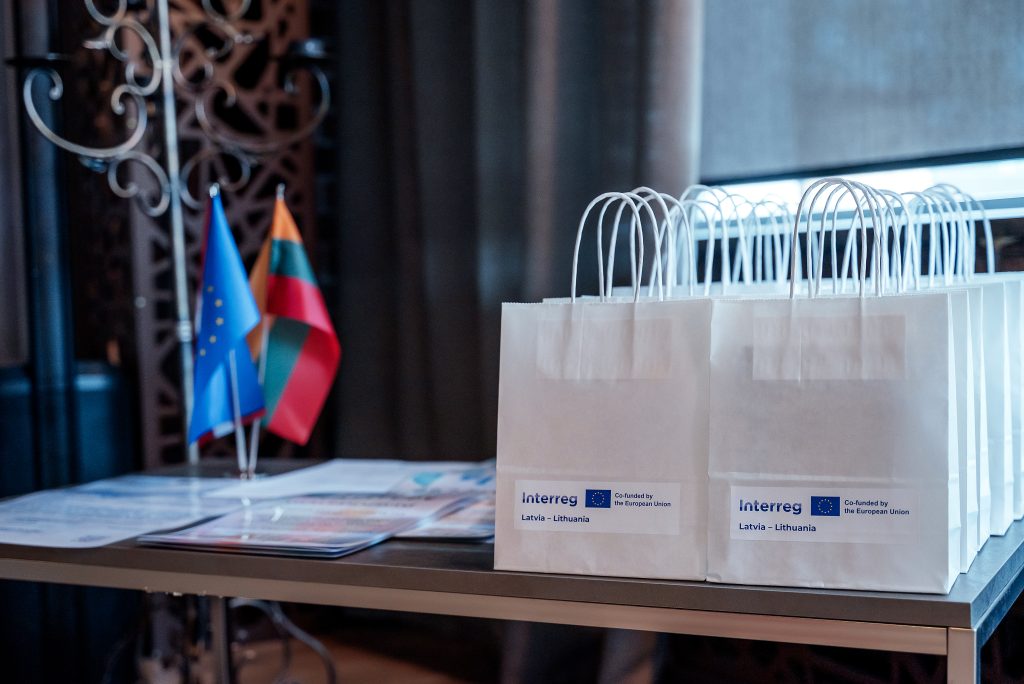 Table displaying white gift bags labeled "Interreg Latvia - Lithuania," alongside flags representing the European Union and Lithuania, at an event promoting cross-border cooperation. Table displaying white gift bags labeled "Interreg Latvia - Lithuania," alongside flags representing the European Union and Lithuania, at an event promoting cross-border cooperation.