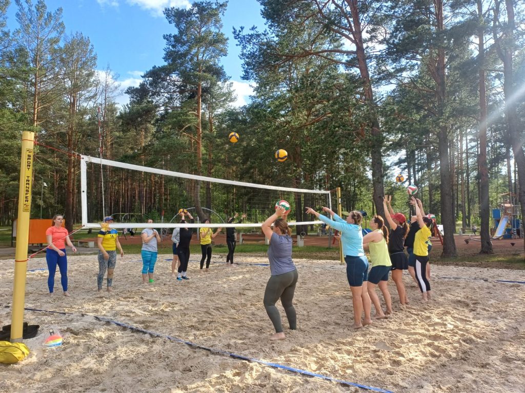 Group of women participating in a beach volleyball practice session on a sandy court surrounded by trees, with a volleyball net and colorful balls in play.