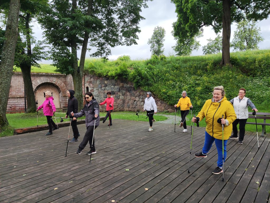 Group of people participating in a Nordic walking session on a wooden platform surrounded by greenery, wearing colorful jackets and using walking poles.