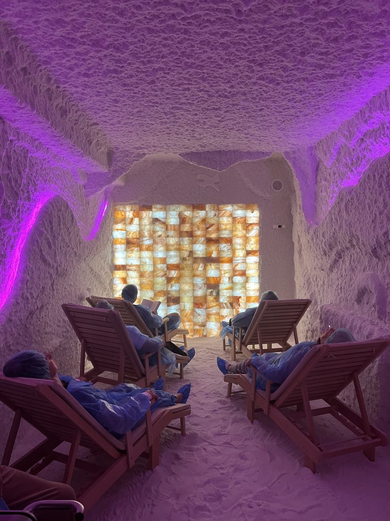 Relaxing salt therapy room with individuals seated on lounge chairs, surrounded by Himalayan salt walls and soft purple lighting, promoting psychosocial and physical rehabilitation for stress relief and social inclusion. Relaxing salt therapy room with individuals seated on lounge chairs, surrounded by Himalayan salt walls and soft purple lighting, promoting psychosocial and physical rehabilitation for stress relief and social inclusion.
