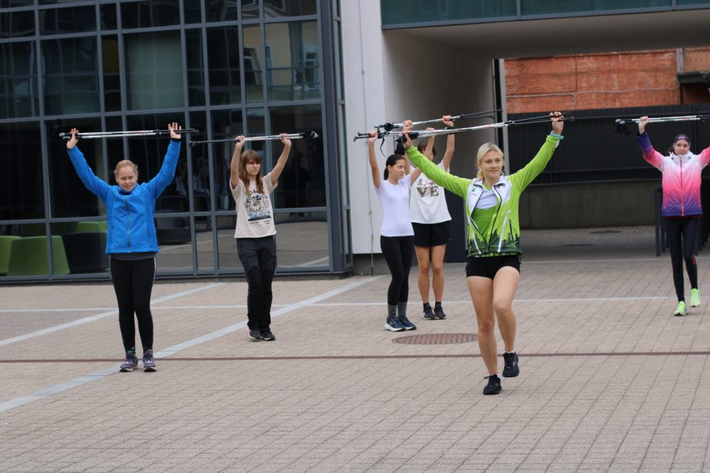 A group of women participating in a fitness class outdoors, using poles for exercise. They are demonstrating various movements with the poles, showcasing teamwork and physical activity. The setting features modern architecture in the background.