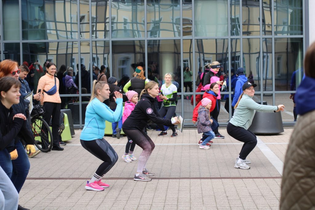 Group of people, including children and adults, participating in an outdoor fitness class, performing squats in front of a modern building with large glass windows. The scene captures a vibrant community exercise event focusing on health and wellness.