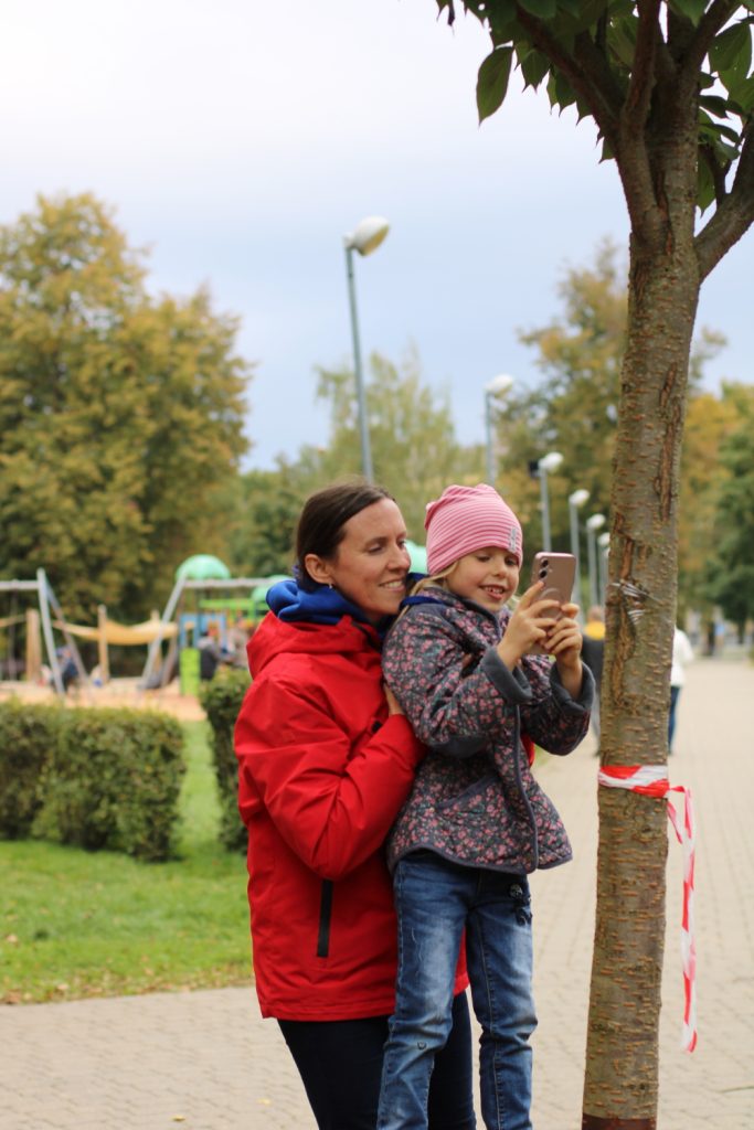 A mother holds her smiling daughter, who is taking a selfie with a smartphone in a park. They are surrounded by trees and playground equipment, showcasing a joyful family outing.