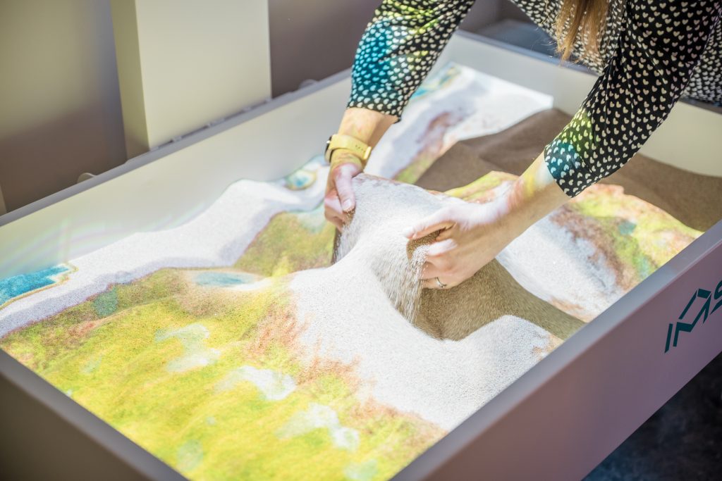 Person manipulating sand in an interactive sand table, showcasing innovative psychosocial rehabilitation methods for better stress management and social inclusion of vulnerable individuals. Person manipulating sand in an interactive sand table, showcasing innovative psychosocial rehabilitation methods for better stress management and social inclusion of vulnerable individuals.
