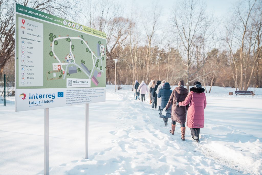 Group of people walking on a snowy path towards a sensory therapy area, with a sign displaying a map and information about the rehabilitation project. The image emphasizes innovative rehabilitation methods for vulnerable individuals, promoting social inclusion. Group of people walking on a snowy path towards a sensory therapy area, with a sign displaying a map and information about the rehabilitation project. The image emphasizes innovative rehabilitation methods for vulnerable individuals, promoting social inclusion.