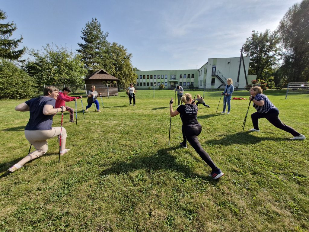 Group of individuals engaging in outdoor Nordic walking exercises on a grassy field, showcasing various lunging techniques with poles. A wooden shed and a building are visible in the background, under a clear blue sky.