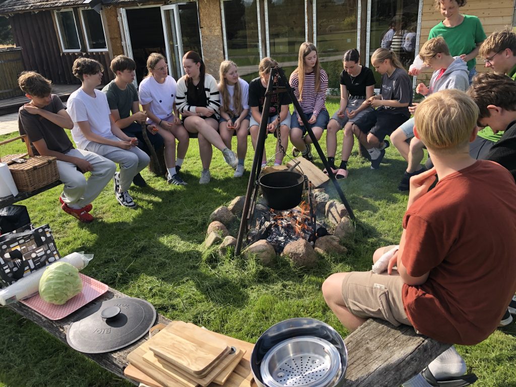 Group of teenagers gathered around a campfire, engaging in cooking activities outdoors. A pot hangs over the fire, with various cooking utensils and fresh ingredients like cabbage visible on a wooden table. The setting features a grassy area and a rustic background, creating a lively atmosphere for socializing and cooking.