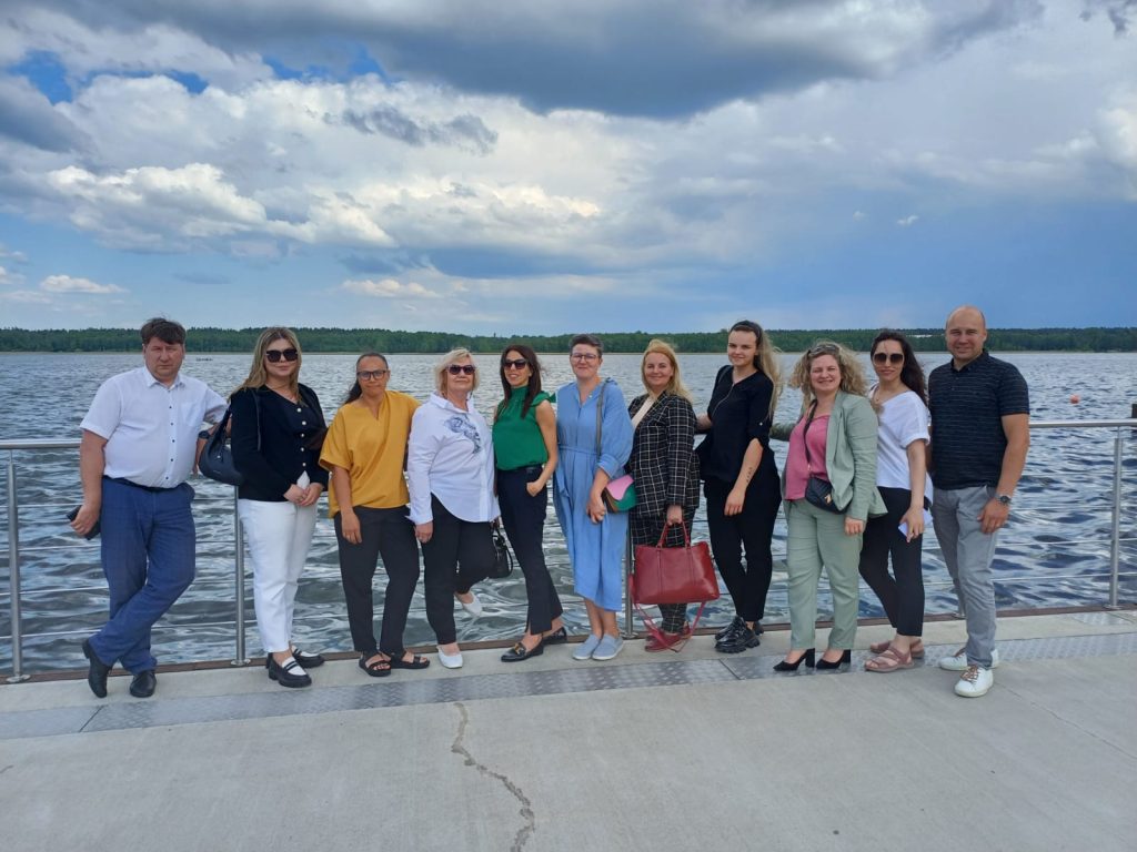 Group of professionals posing together by a lake, showcasing a variety of outfits against a scenic backdrop of water and clouds. The image captures a diverse team enjoying a day outdoors, highlighting collaboration and camaraderie.
