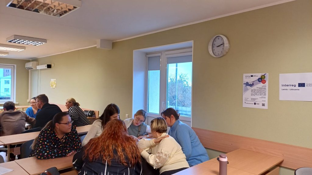 Group of people collaborating in a classroom setting, engaged in discussions and teamwork. The room features a clock, windows with natural light, and educational materials on the walls, highlighting an interactive learning environment.