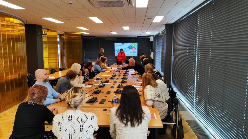 Meeting in a modern conference room with attendees engaged in discussion, featuring a presenter in a red jacket and a large screen displaying information. The room is equipped with audio-visual technology and has large windows with blinds.