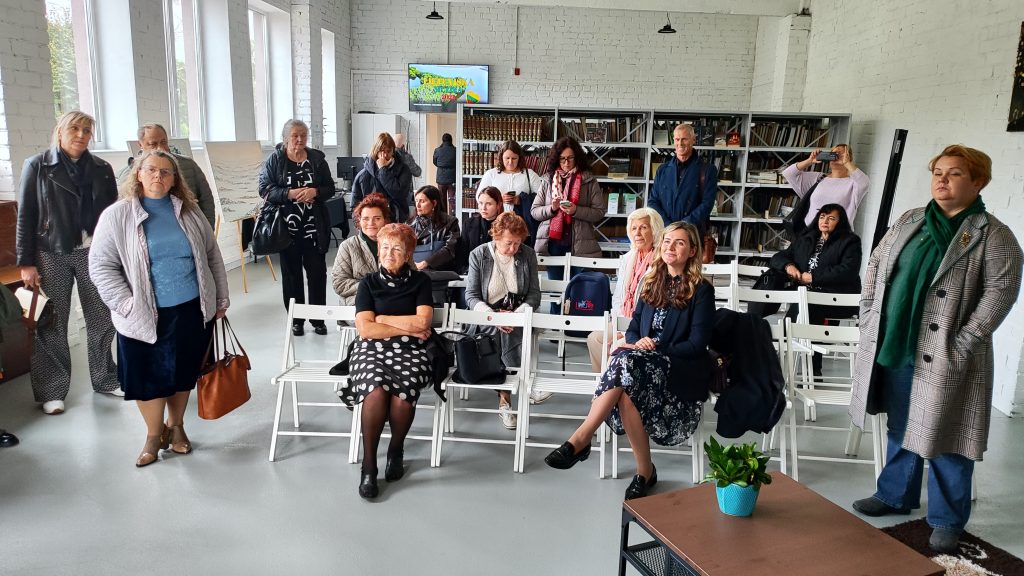 A diverse group of people attending a community event in a bright, modern space, featuring a mix of ages and styles, seated and standing, with bookshelves and a screen displaying information in the background.