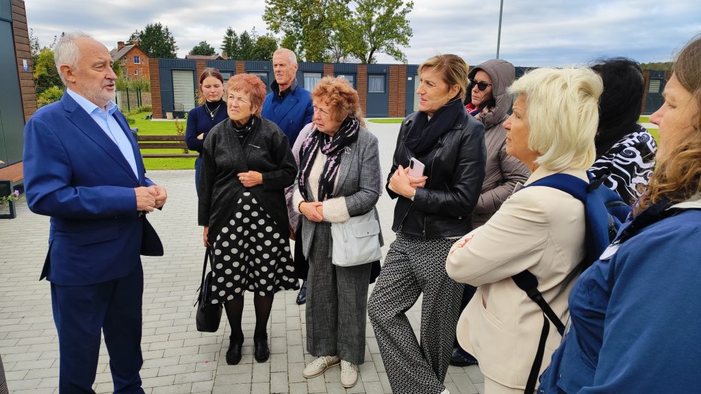 Group of people listening attentively to a speaker in a blue suit during an outdoor event, surrounded by greenery and modern architecture. The audience includes men and women of various ages, engaged in discussion.
