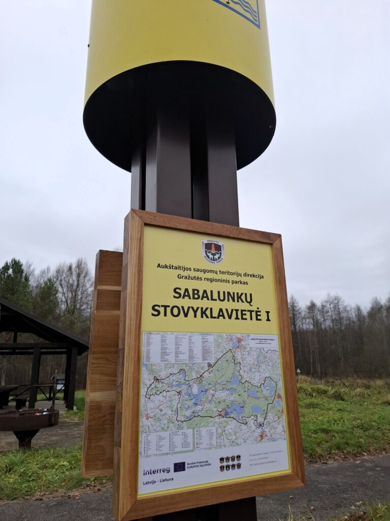 Signpost for Sabalunki Stovyklavietė 5, featuring a detailed map and information about the camping area, surrounded by a forested landscape.