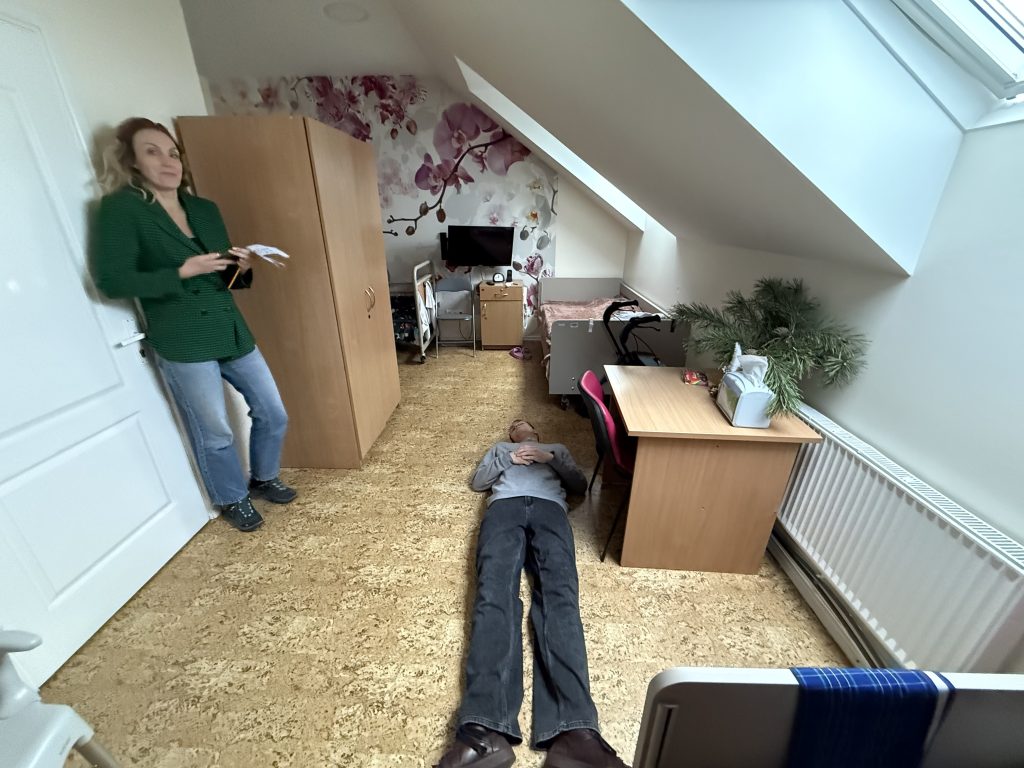 A woman standing in a room with wooden flooring, gesturing towards a person lying on the ground. The room features a couch, a desk, and decorative elements on the walls, indicating a cozy living space.