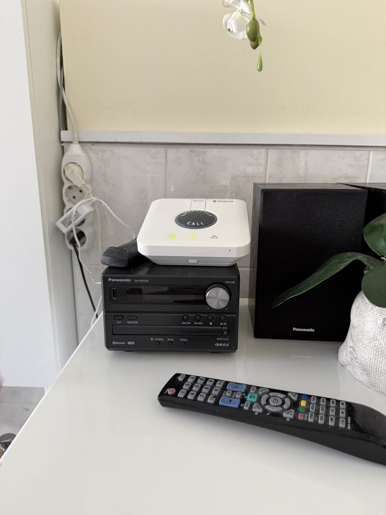 Compact audio system with a white speaker on top, a black base unit, and a remote control on a white table, alongside a decorative plant.