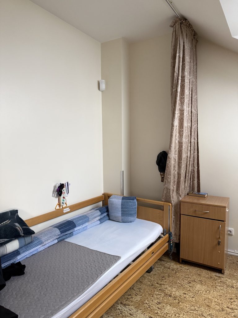 Cozy bedroom interior featuring a single bed with blue and gray bedding, a wooden nightstand, and a window with curtains. The room is well-lit and designed for comfort.